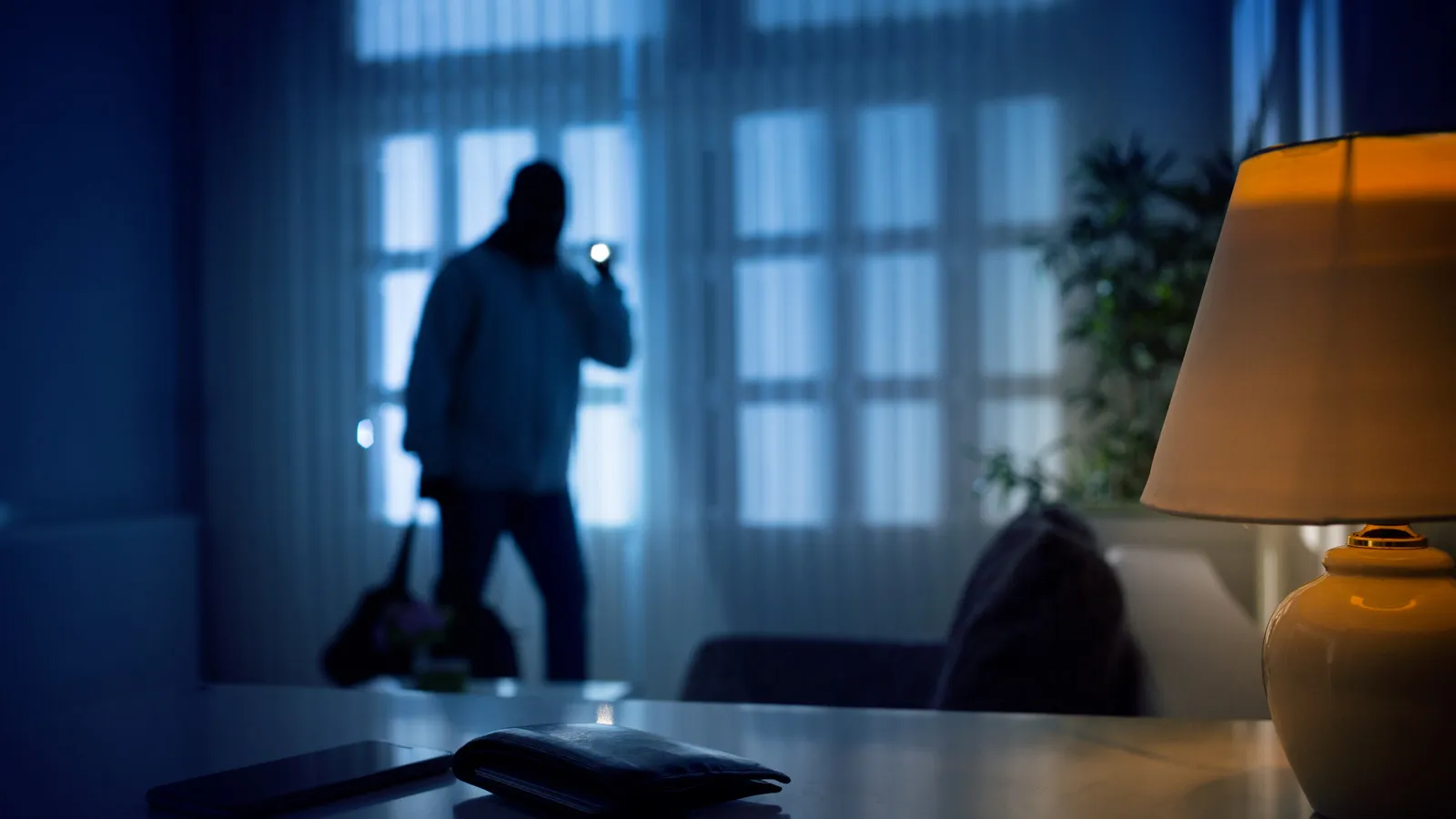 burglar in a dark room with face covered and flashlight looking at a wallet and phone
